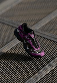 Black and purple athletic shoe with a mesh upper, contrasting silver accents, and a textured sole. Features elastic lacing for secure fit.