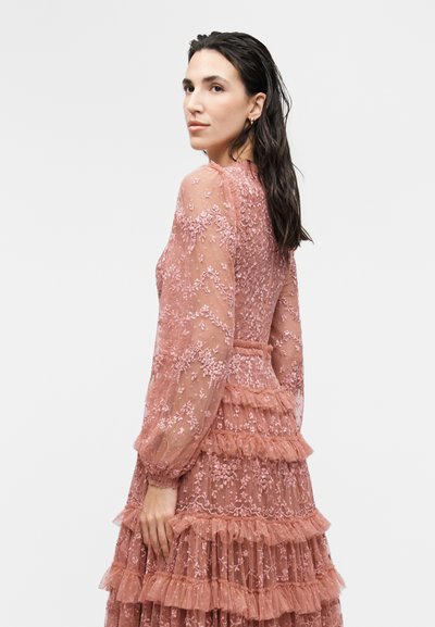 Γυναίκα με σκούρα βρεγμένα μαλλιά, που φοράει μακριμάνικο διάφανο ροζ δαντελένιο φόρεμα με κεντήματα λουλουδιών και στρωτές βολάν, σε προφίλ.