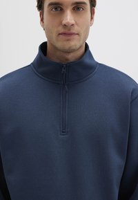 Dunkelblauer Pullover mit einem Viertel-Reißverschluss-Design, hergestellt aus strukturiertem Stoff. Verfügt über einen hohen Kragen und minimalistische Nähte.