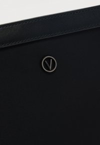 Mustan kankaan ja nahan laukun yksityiskohta, jossa pieni metallinen pyöreä logo, jossa on tyylitelty "V", keskitetty ommellun nahkareunan alapuolelle.