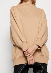 Beige översize sweatshirt med hög krage, ribbade muddar och fåll. Har en liten vit Nike-logotyp på bröstet. Slät textur.