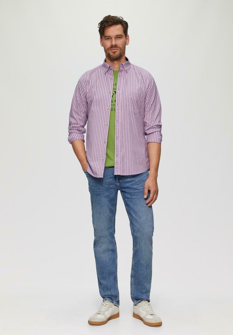 Heren overhemd met kraag in licht paars met witte strepen, gelaagd over een groene T-shirt met grafische print; gecombineerd met blauwe spijkerbroek en witte sneakers.