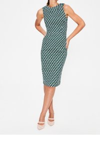 Grön och vit mönstrad ärmlös klänning med en figurnära silhuett, knälång vidd och slät tygtextur. Bärs med ljusa klackar.