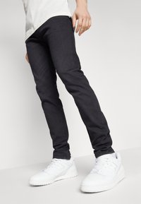 Svarta slim-fit jeans med en slät yta, kombinerat med vita sportiga sneakers med tjock sula och logo-detalj på sidan.