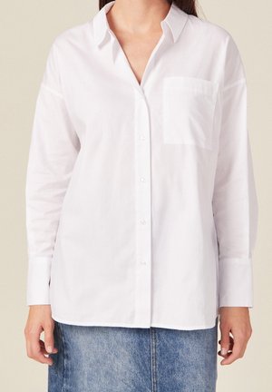 Frau trägt ein weißes Hemd mit langen Ärmeln und Brusttasche sowie einen blauen Jeansrock und steht vor einem neutralen Hintergrund.