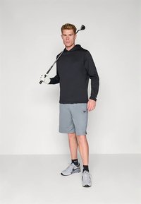 Μαύρο φούτερ με μακριά μανίκια, γκρι σορτς και γκρι αθλητικά παπούτσια. Κρατάει ένα μπαστούνι γκολφ, φορώντας άσπρο γάντι γκολφ. Απλός, λειτουργικός αθλητικός σχεδιασμός.