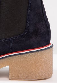 Botín de ante azul marino con un panel elástico negro texturizado, detalle de rayas blancas y rojas en la suela, y un tacón de plataforma beige grueso.