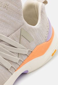 Scarpa atletica beige con una tomaia in tessuto testurizzato, lacci elastici e accenti colorati arancioni e viola sulla suola.