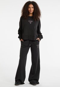 Set di felpa nera e pantaloni tuta con logo "GUESS" in rosa, dalla vestibilità rilassata. Tessuto morbido, maniche lunghe e pantaloni a gamba ampia.