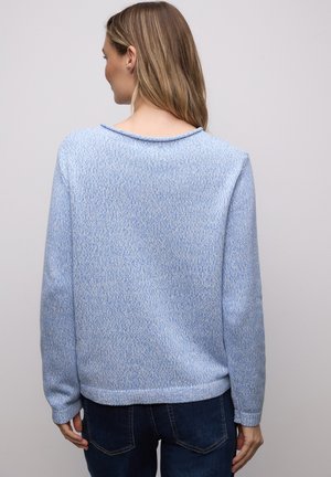 Femme aux cheveux longs portant un pull en maille texturée bleu clair et un jean bleu foncé, vue de dos sur un fond clair uni.