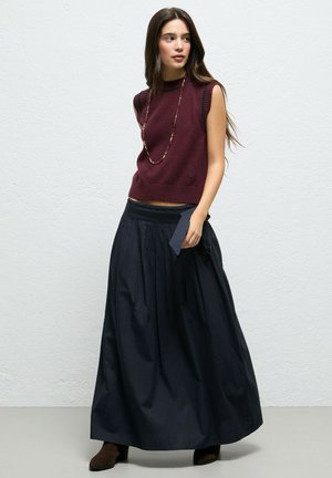 Mujer que lleva un suéter sin mangas color granate, collar largo, falda larga azul marino y botines oscuros, de pie frente a una pared blanca lisa.