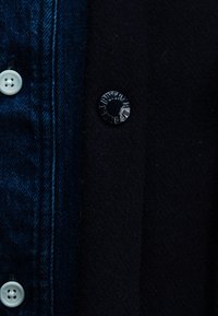 Denim jacket with a dark blue collar, featuring a large black button and a prominent white button. The fabric has a smooth texture.