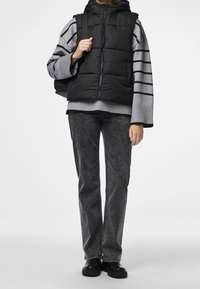 Svart pufferväst med huva över en grårandig tröja, tillsammans med mörkgrå jeans och svarta stövlar. Texturerade material, avslappnad design.