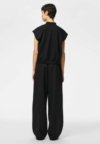 Osoba s kratkom tamnom kosom stoji okrenuta leđima, nosi crnu majicu bez rukava i široke crne hlače sa sitnim prugama.