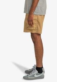 Khaki shorts with a relaxed fit, made from cotton. Features side pockets and snap closures. Paired with gray sneakers and black socks.