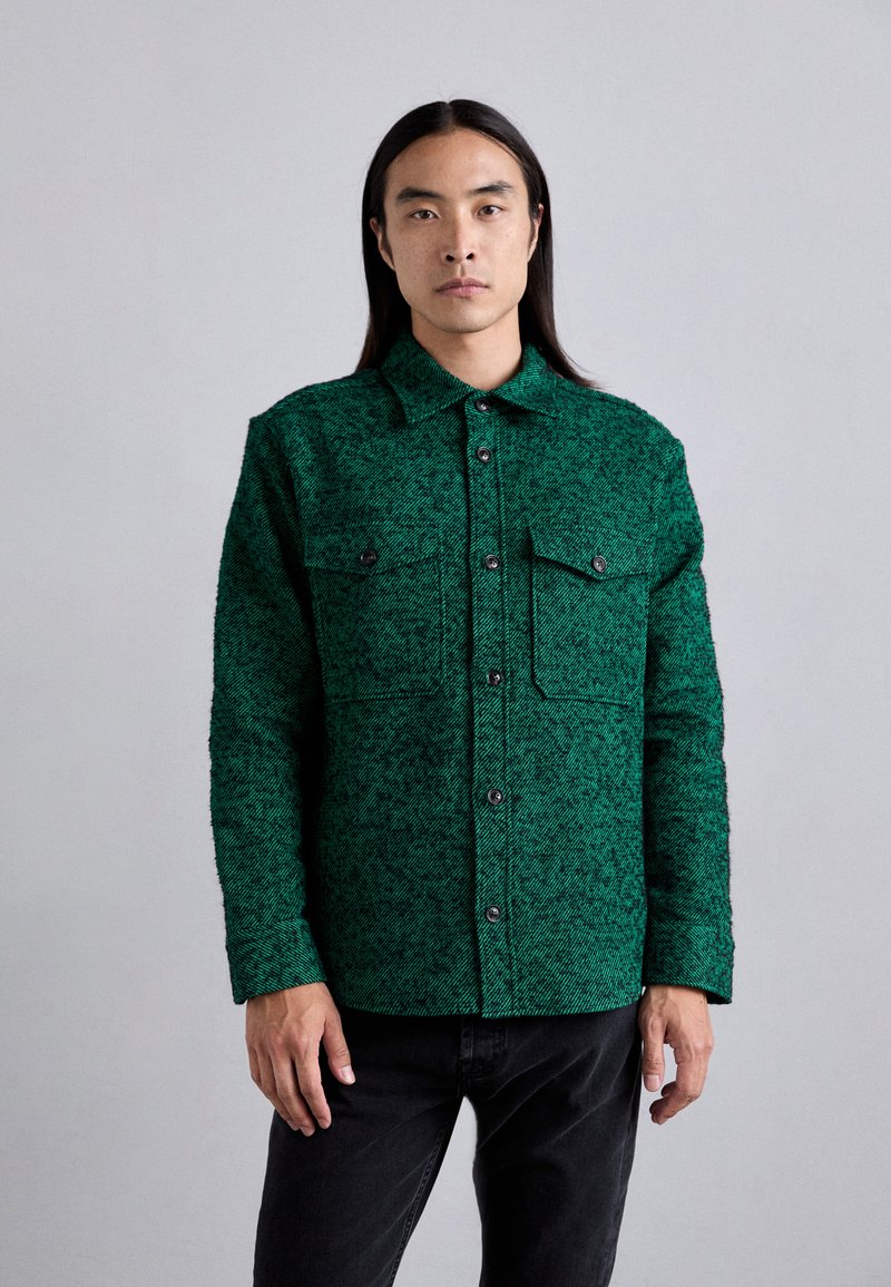 Chemise boutonnée verte, texturée, avec deux poches poitrine et un col, ornée de boutons noirs et d'un motif tissé unique.