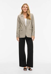 Femme debout portant un blazer beige, un haut blanc, un pantalon noir à jambes larges et des talons ouverts noirs, souriant avec les mains détendues le long du corps.