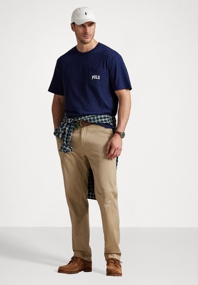 Granatowa bawełniana koszulka z logo "POLO", khaki spodnie, koszula w kratę przewiązana w talii, brązowe buty i biała czapka. Codzienny strój z wyraźnymi warstwami.