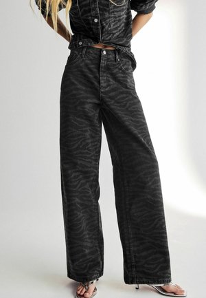 Personne portant un jean noir taille haute à jambes larges avec un motif zèbre subtil et une veste courte assortie, associée à des sandales à talons argentées.