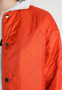 Pikowana pomarańczowa kurtka o gładkiej teksturze, z zatrzaskami oraz szarym kapturem pod spodem, podkreślająca warstwowy styl.