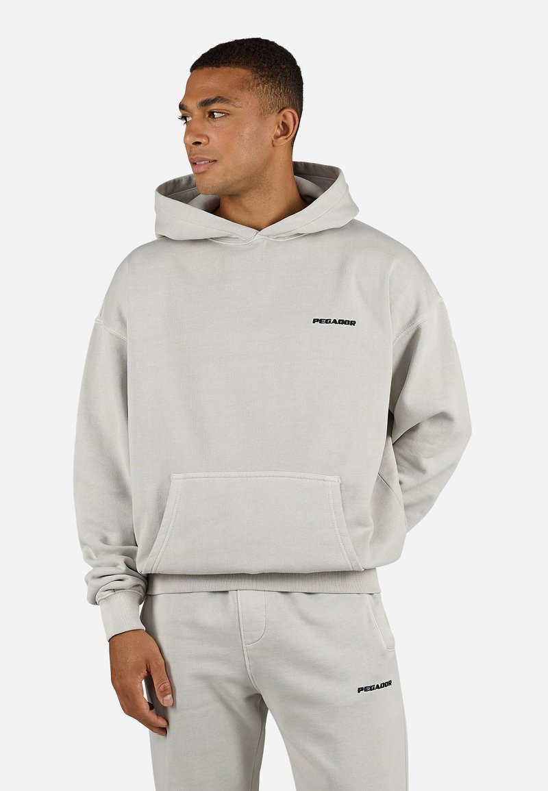 Lichtgrijze hoodie van zacht materiaal, met een voorzak en het "PEGADOR" logo op de borst in zwart. Ruime pasvorm met ribgebreide manchetten.