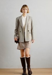 Blazer à carreaux beige et gris, associé à un haut léger et des bottes marron jusqu'au genou avec un bord en fourrure. Le modèle se tient sur un sol en bois clair.