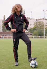 Chándal atlético negro con acentos rojos, de pie sobre un balón de fútbol, en un campo de hierba. Footwear presenta un diseño elegante y un patrón de agarre.