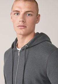 Sudadera gris con cremallera hecha de una mezcla de algodón. Presenta una capucha con cordón, cremallera frontal y detalles de canalé en los puños y el dobladillo.