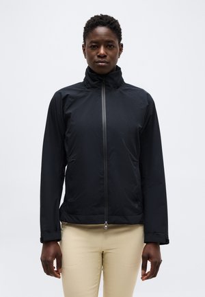 Suaugęs trumpais plaukais, dėvintis juodą striukę su užtrauktuku ir smėlio spalvos kelnes, stovintis prieš paprastą šviesią foną.