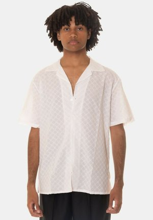 Camicia bianca a maniche corte con motivo a rombi, dotata di colletto a punte e chiusura con bottoni sul davanti. Tessuto leggero e strutturato.