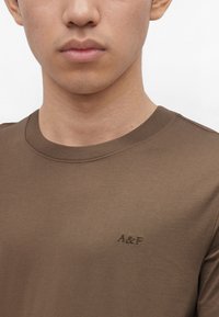 Νέος άνδρας με καφέ T-shirt με στρογγυλή λαιμόκοψη και κέντημα «A&F» στο αριστερό στήθος, σε απλό λευκό φόντο.