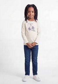 Νεαρή κοπέλα με πλεξούδες που φοράει κρεμ μπλούζα μακρυμάνικη με σχέδιο κερασιού και το κείμενο «hello cute», μπλε τζιν και λευκά αθλητικά παπούτσια.