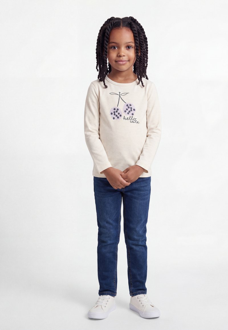 Νεαρή κοπέλα με πλεξούδες που φοράει κρεμ μπλούζα μακρυμάνικη με σχέδιο κερασιού και το κείμενο «hello cute», μπλε τζιν και λευκά αθλητικά παπούτσια.