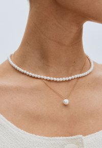 Zwei geschichtete Halsketten: eine mit weißen Perlen und die andere mit einem einzelnen Perlenanhänger an einer goldenen Kette, die auf nackter Haut getragen wird.