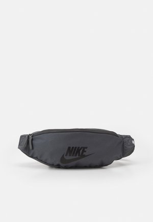 Banane noire Nike avec fermeture éclair et logo sur le devant, posée sur un fond gris clair uni.