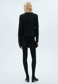 Blazer negru croit, cu accente din nasturi, fustă neagră cu volane în straturi, ciorapi negri opaci și pantofi cu toc gros. Vedere din spate.