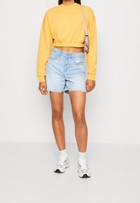 Felpa gialla corta, pantaloni shorts in denim blu chiaro con orlo sfrangiato, calzini bianchi e sneakers bianche con dettagli grigi. Borsa a piccolo motivo.