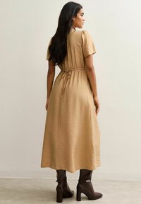 Longue robe beige à manches courtes, taille élastique et tissu texturé. Complétée par des bottes à talons marron hautes.