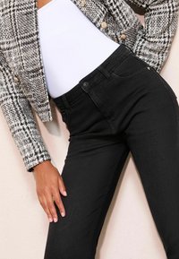 Fekete, magas derékú, slim fit farmernadrág, elülső gombos záródással és lapos elülső zsebekkel. Fehér felsővel és mintás blézerral viselve.