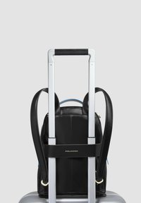 Mochila de cuero negro sujeta al asa de una maleta plateada. Cuenta con dos correas para los hombros y cierre con cremallera. Diseño minimalista; logo visible.