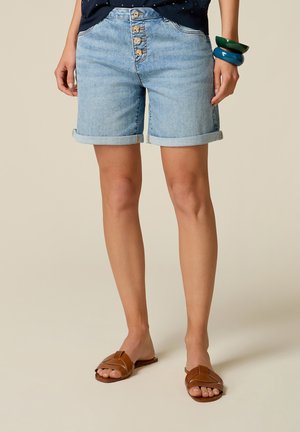 Persoon die lichtblauwe opgerolde denimshorts met vier knopen en bruine sandalen met open tenen draagt tegen een neutrale achtergrond.