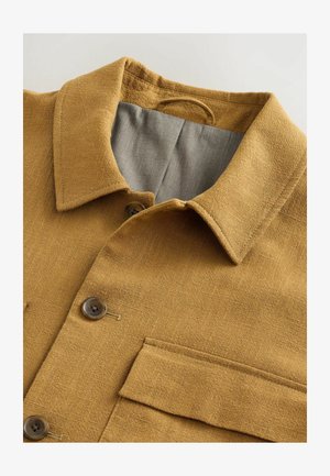 Ovrig skjorte i gyldenbrun tekstureret stof med klassisk krave, grå foring, to knapper og en brystlomme.