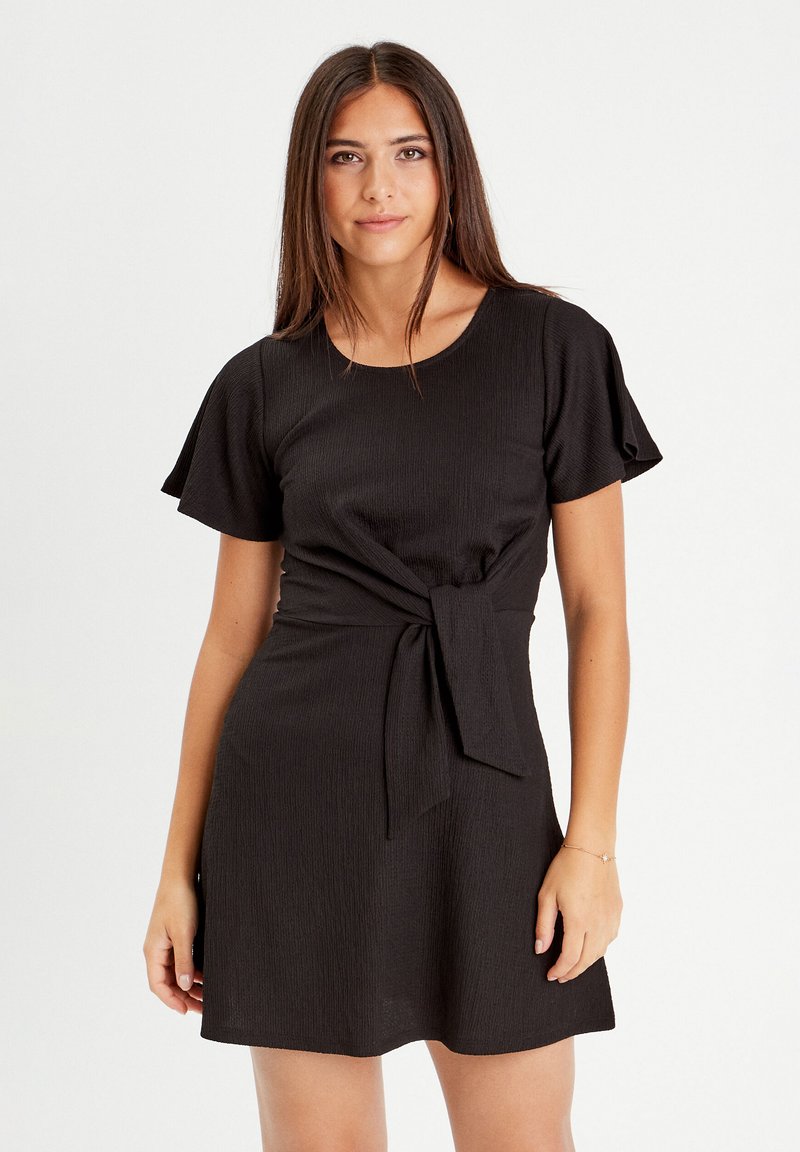 Robe noire en tissu texturé, avec des manches courtes volantes, un col rond et un détail noué à la taille.