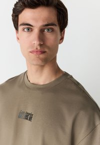 Olivgrünes Baumwoll-T-Shirt mit Rundhalsausschnitt und kurzen Ärmeln, mit schwarzem Text "Los Angeles MELROSE AVE" auf der Brust. Glatte Textur.