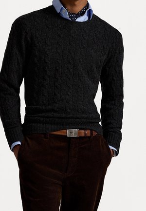 Mann trägt schwarzen Strickpullover mit Kabelmuster über blauem Hemd mit Kragen und gepunktetem Ascot-Schal, braune Cordhose und braunen Ledergürtel, Hände in den Taschen.