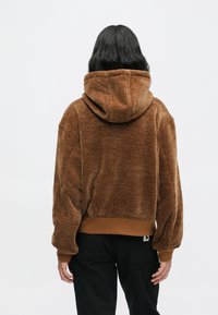 Pessoa com cabelo longo e escuro veste um hoodie texturado castanho e calças pretas, de pé com as costas viradas para o espectador, contra um fundo plano.