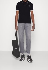 Man i svart polo shirt och grå jeans som håller en svart tote-väska med text, iförd svarta och vita sneakers mot en enhetlig bakgrund.