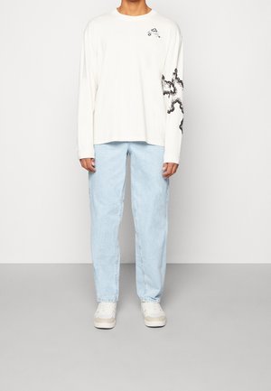 Personne portant un t-shirt à manches longues blanc cassé avec un graphique noir sur la manche et un jeans bleu clair, se tenant devant un fond gris clair uni.