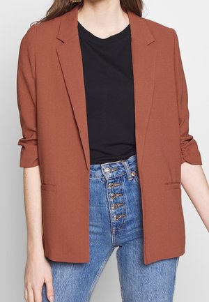 Frau trägt einen rostfarbenen Blazer mit gerafften Ärmeln über einem schwarzen Hemd und hoch taillierte blaue Jeans mit mehreren Knöpfen.
