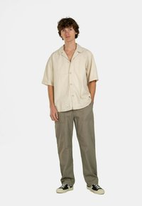 Helles beige Hemd mit kurzen Ärmeln und lockerer Passform, mit einer Knopfleiste. Kombiniert mit locker sitzenden grauen Hosen und schwarzen Sneakers.